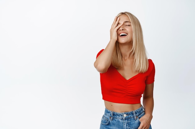 Attactive blond woman in stylish summer outfit laughing and touching her face smiling carefree standing over white background
