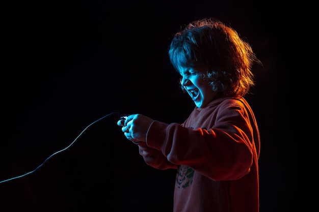 Attented playing videogames. Caucasian boy's portrait on dark studio background in neon light. Beautiful curly model. Concept of human emotions, facial expression, sales, ad, modern tech, gadgets.