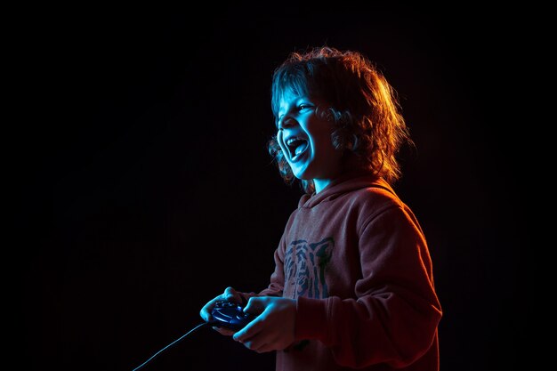 Attented playing videogames. Caucasian boy's portrait on dark studio background in neon light. Beautiful curly model. Concept of human emotions, facial expression, sales, ad, modern tech, gadgets.