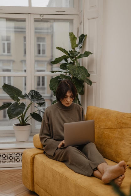 Attentive adult caucasian woman reads message ebook or information on her notebook while sitting on yellow sofa in spacious room