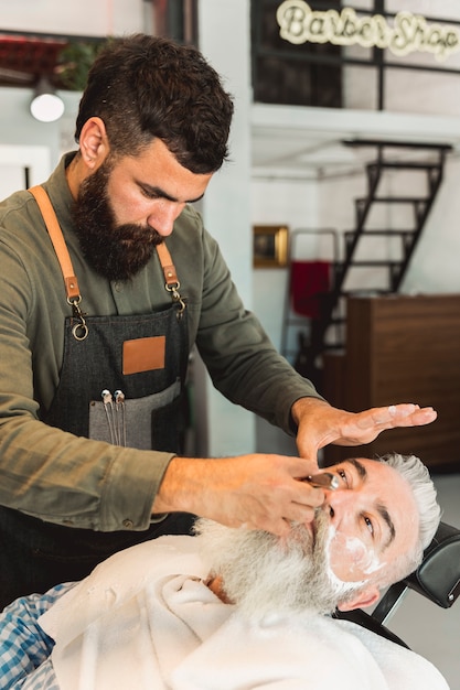 Foto gratuita barbiere attento che rade barba al cliente nel negozio di barbiere