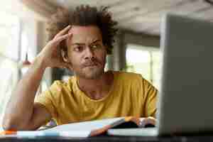 Free photo attentive black young male sitting indoors in front of opened laptop being very serious while reading scientific article online trying to find main point of it and to write review on this topic