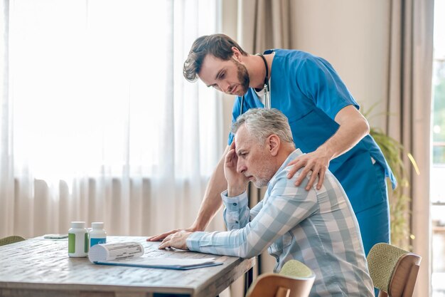 Attentive focused bearded young Caucasian male doctor with a stethoscope bending over an upset man