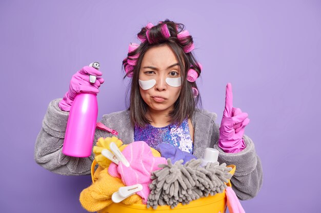 Attentive housewife applies hair rollers for making hairstyle wears beauty patches under eyes dressed in domestic robe rubber gloves cares about purity holds detergent poses near laundry basket