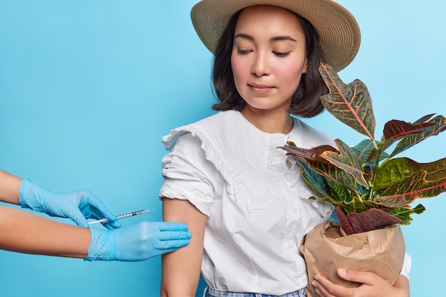 Free photo attracitve asian lady with dark hair holds potted houseplant gets vaccine in arm to protect herself from coronavirus wears white blouse fedora isolated over blue wall