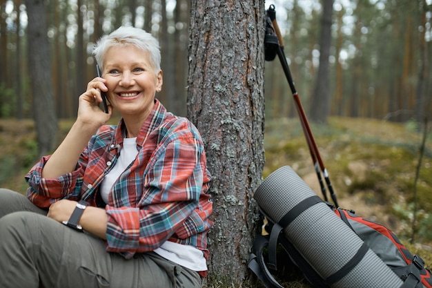 無料写真 森でハイキング、木の下に座って、携帯電話で友人と話している間、小さな休憩を持っている格子縞のシャツの魅力的なアクティブな女性年金受給者。電話をかける中年女性旅行者