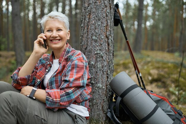 모바일에서 친구에게 말하기, 숲에서 하이킹하는 동안 작은 휴식을 취하는 격자 무늬 셔츠에 매력적인 활성 여성 연금 수령자. 중간 나이 든된 여자 여행자 전화 통화