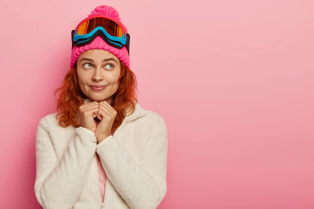 Attractive active woman has dreamy expression,  wears pink hat with ski goggles, copy space