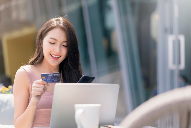 Привлекательная взрослая азиатская женщина делает покупки в Интернете с помощью ноутбука, держащего кредитную карту и смартфон для покупки продукта на фоне концепции концепции бизнес-идей для сада на открытом воздухе