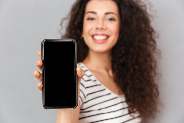 Attractive adult girl with ring in nose demonstrating her mobile phone advertising new model being glad while isolated against grey wall