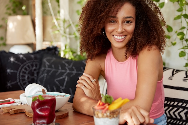 Attractive African American dark skinned woman dressed in pink t shirt, sits crossed hands at table in cafe, surrounded with cocktails and sweet dessert, has pleased expression, has good rest.