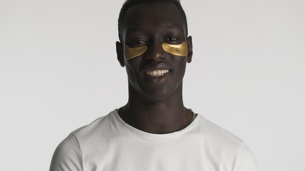 Привлекательный афроамериканец с золотыми повязками на глазах улыбается в камеру на белом фоне. Уход за лицом