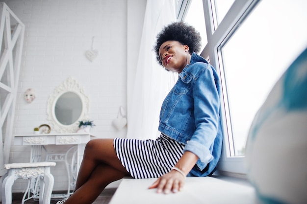 Attraente donna afroamericana con capelli afro su gonna e giacca di jeans poste in camera bianca modello nero alla moda