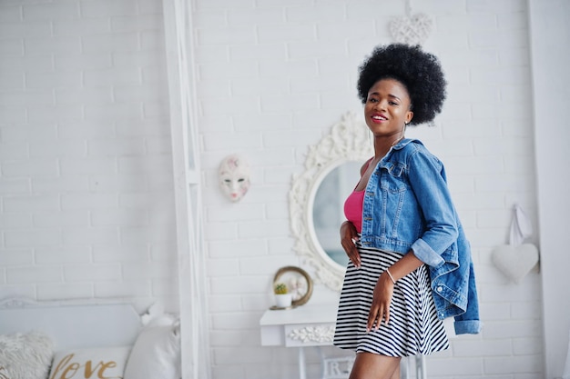 Attractive african american woman with afro hair wear on skirt and jeans jacket posed at white room Fashionable black model