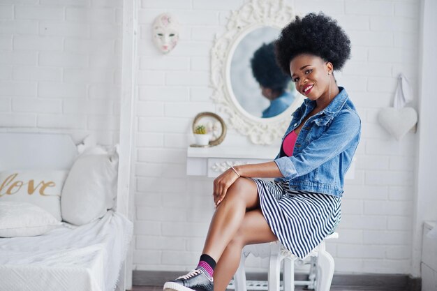 Attractive african american woman with afro hair wear on skirt and jeans jacket posed at white room Fashionable black model