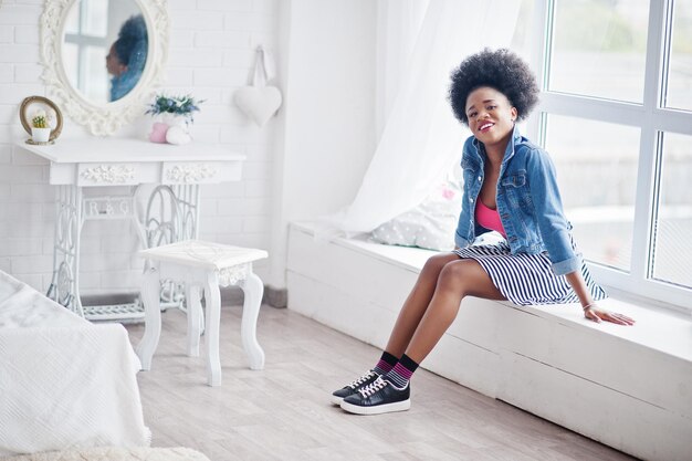 Attractive african american woman with afro hair wear on skirt and jeans jacket posed at white room Fashionable black model