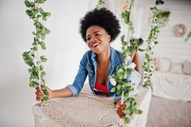 Attractive african american woman with afro hair wear on skirt and jeans jacket posed at white room on swing Fashionable black model