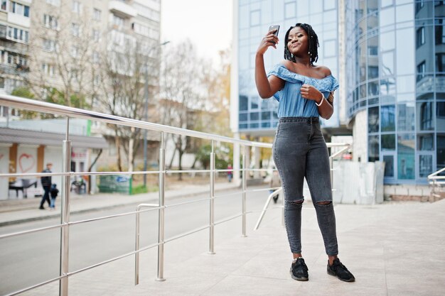 Attractive african american woman with dreads in jeans wear posed near railings against modern multistory building making selfie at mobile phone