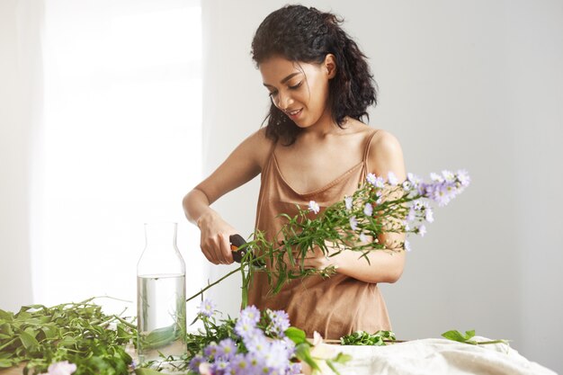 Attractive african female florist cutting stems at workplace. White wall.