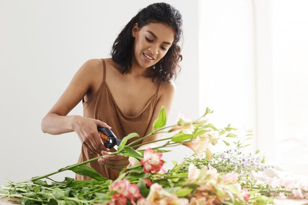 Attractive african female florist smiling cutting stems working in flower shop over white wall.