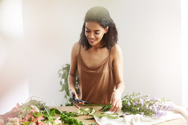 Attractive african female florist smiling cutting stems working in flower shop over white wall.