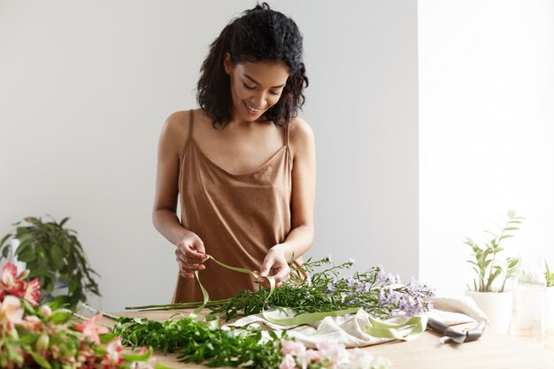 Attractive african female florist smiling holding green ribbon working with flowers at workplace over white wall.