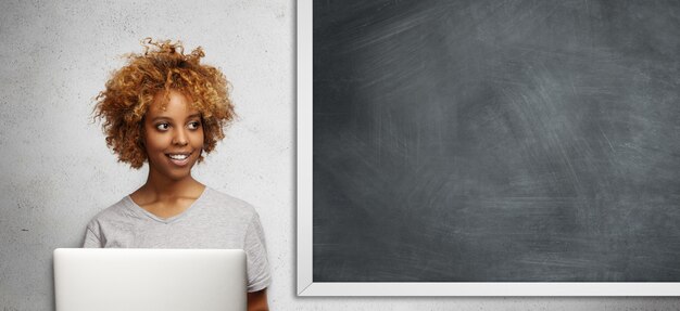 Attractive African student with Afro hairstyle and cute smile, looking aside with pensive expression, using free Internet connection on laptop computer, doing classwork, sitting at blackboard