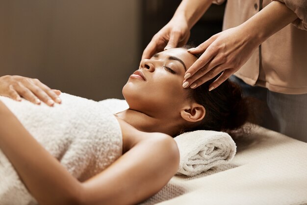 Attractive african woman enjoying face massage in spa salon.