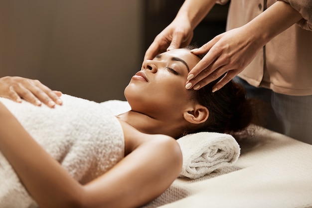 Attractive african woman enjoying face massage in spa salon.
