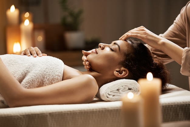 Attractive african woman enjoying face massage in spa salon.