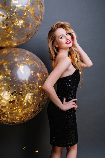 Attractive amazing woman from back in luxury black dress, with long curly blonde hair, red lips smiling with big balloons full with golden tinsels. Celebrating, party.