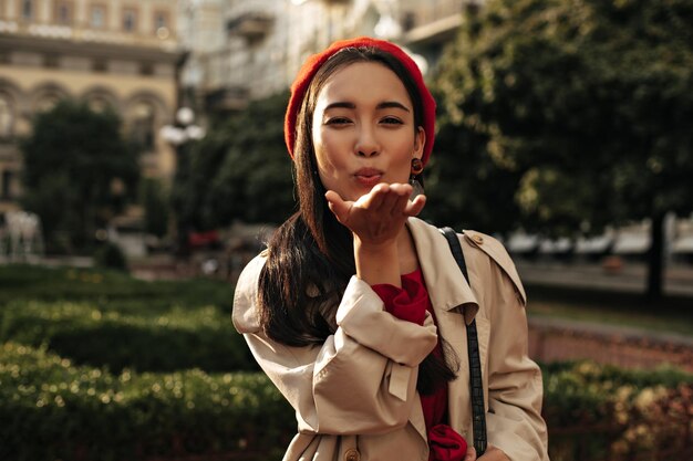 Attractive Asian brunette woman in red beret bright dress and stylish beige trench coat blows kiss and poses in good mood outside