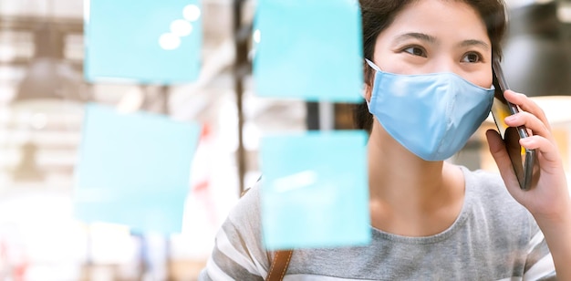 Привлекательная азиатская женщина в темно-синем свитере, работающая в защитной вирусной маске, работающая в совместном рабочем пространстве с пластиковой перегородкой, социальное дистанцирование для сиденья, новый нормальный образ жизни