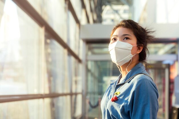 Attractive asian female woman casual cloth wearing face mask protection standing next to big glasses modern frame window with sun light flare subset moment new normal lifestyle healthcare concept