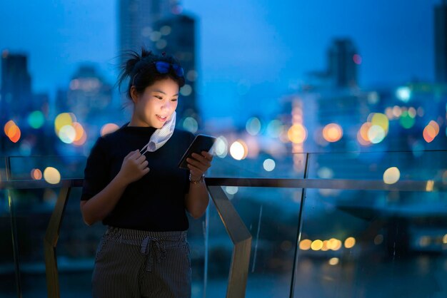 Привлекательная азиатская женщина носит защитную маску, ручное использование смартфона, технология связи на фоне боке, светлое городское здание, фон, концепция бизнес-идей