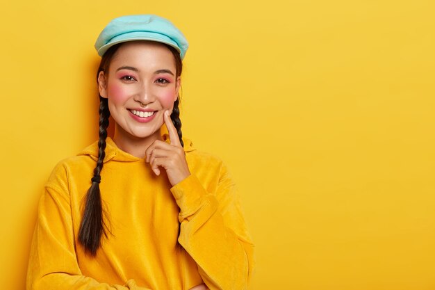 ピンクのメイクで魅力的なアジアの女性、頬に人差し指を保ち、良いアイデアを熟考し、フード付きのカジュアルな黄色のベルベットのスウェットシャツを着て、優しく微笑む