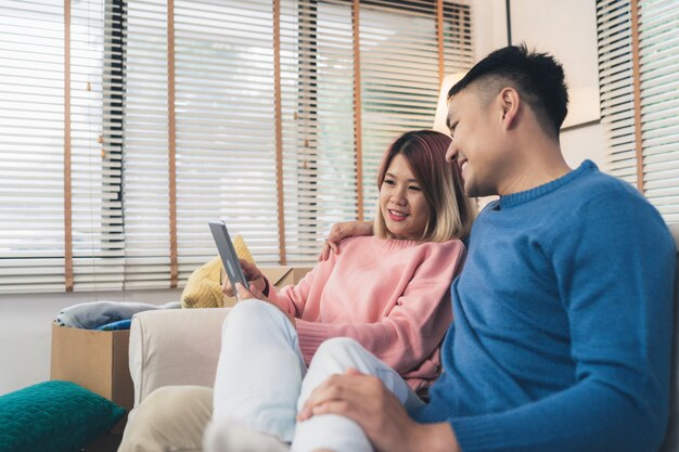 Attractive Asian sweet couple using tablet while lying on the sofa when relax in their living room 