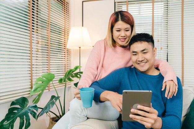 Attractive Asian sweet couple using tablet while lying on the sofa when relax in their living room