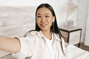 Free photo attractive asian woman in beige plaid jacket and white tshirt smiles takes selfie in bedroom and listens to music in wireless headphones