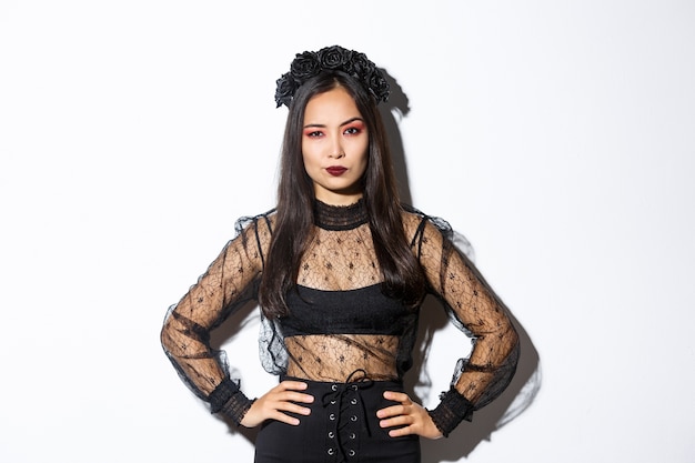 Free photo attractive asian woman in halloween costume looking disappointed and skeptical. female in black lace dress and wreath looking arrogant, trick or treat in witch outfit, standing white background.