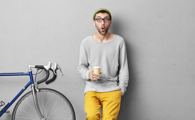 Привлекательный удивленный мужчина, продающий спортивный велосипед, пьющий кофе из чашки на вынос, с огромным удивлением смотрит на то, как замечает множество покупателей. Небритый мужчина после остановки на велосипеде на улице