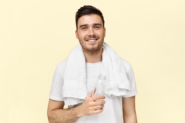 Attractive athletic young male with towel around his neck enjoying fresh drinking water, holding bottle, resting after training at gym