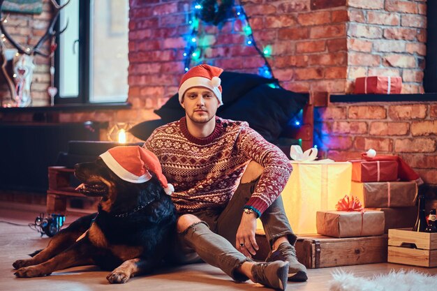 Привлекательный бородатый хипстер сидит на полу со своей собакой-ротвейлером в комнате с рождественским украшением.
