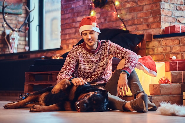 Attractive bearded hipster male sits on a floor with his Rottweiler dog in a room with Christmas decoration.