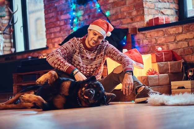수염을 기른 매력적인 힙스터 남성은 크리스마스 장식이 있는 방에서 로트와일러 개와 함께 바닥에 앉아 있습니다.