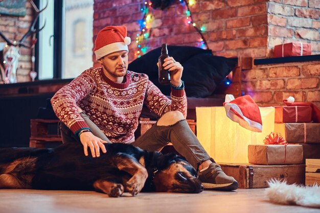 魅力的なあごひげを生やしたヒップスターの男性は、クリスマスの装飾が施された部屋で彼のロットワイラー犬と一緒に床に座っています。