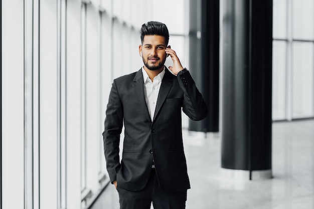 An attractive bearded Indian businessman in a modern office center, wearing a black suit, holds his hand near a wireless earphone