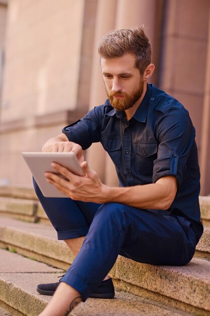 The attractive bearded male sits on a step and using a tablet PC in a city.