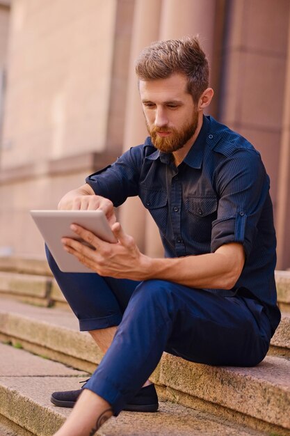 Привлекательный бородатый мужчина сидит на ступеньке и пользуется планшетным ПК в городе.