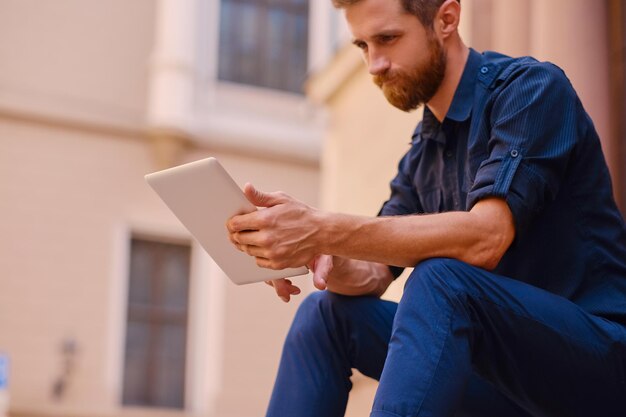 The attractive bearded male sits on a step and using a tablet PC in a city.